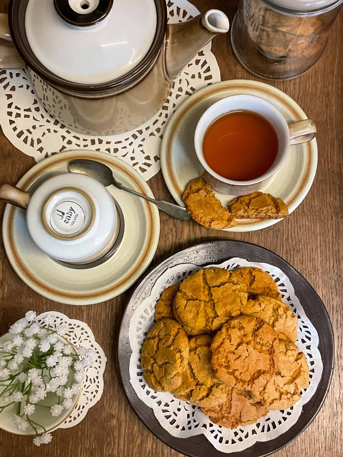 Tea table setting with cup and saucer, teapot, fairings on a plate, and a small dish of flowers.