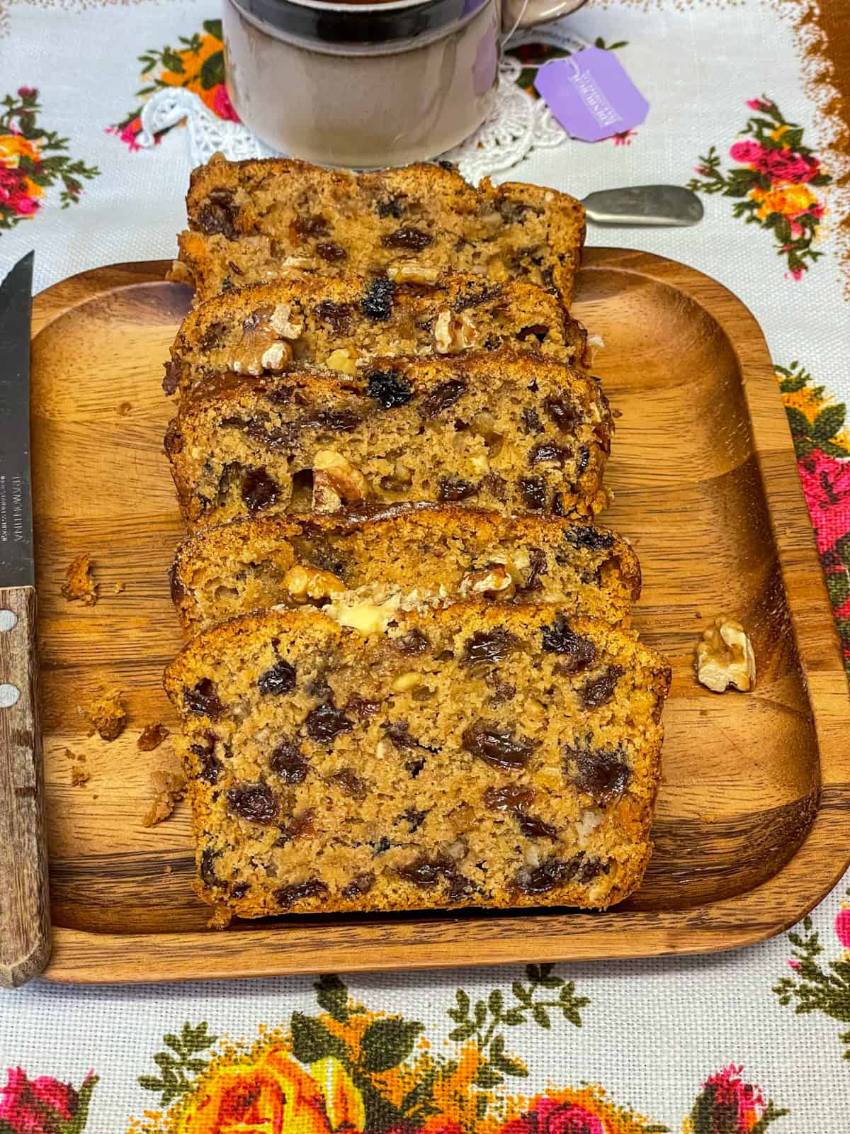 Slices of country tea bread on a wooden board with a flowered patterned table cloth, tea cup in background.