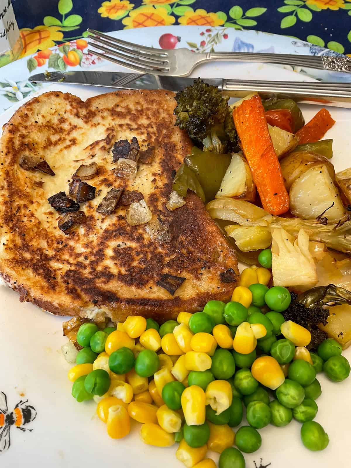 Victorian bread steaks served on a plate with roast mixed vegetables and peas and corn, silver fork and knife in background.