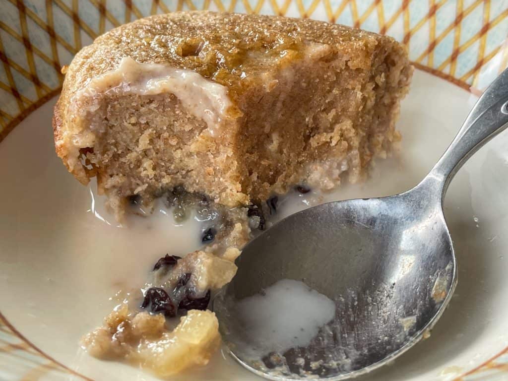 A slice of wheatmeal pudding in serving bowl with cream and silver spoon.
