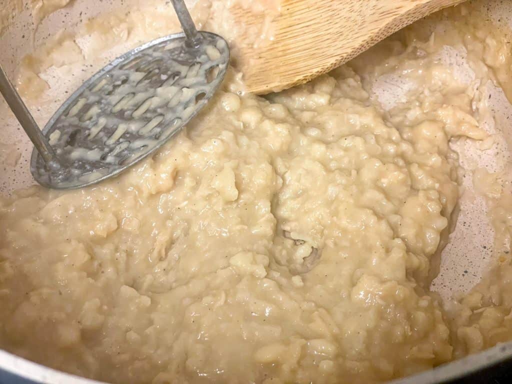 White beans mashed with potato masher in saucepan.